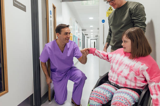 A support worker in scrubs fist-bumping a girl in a wheelchair in a hallway, with her father alongside