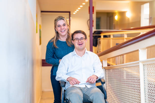 A support worker standing behind a smiling young man in his wheelchair in a bright indoor hallway