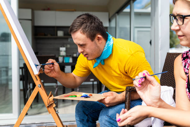 A young man painting on an easel outdoors while a support worker joins in beside him