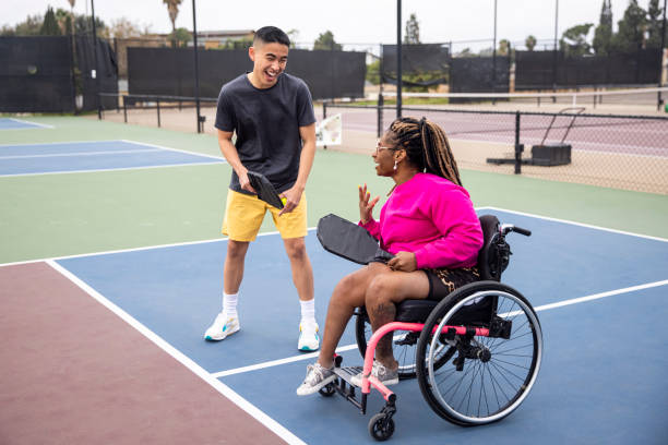 A young man and a woman in a wheelchair laughing together while playing pickleball on an outdoor court